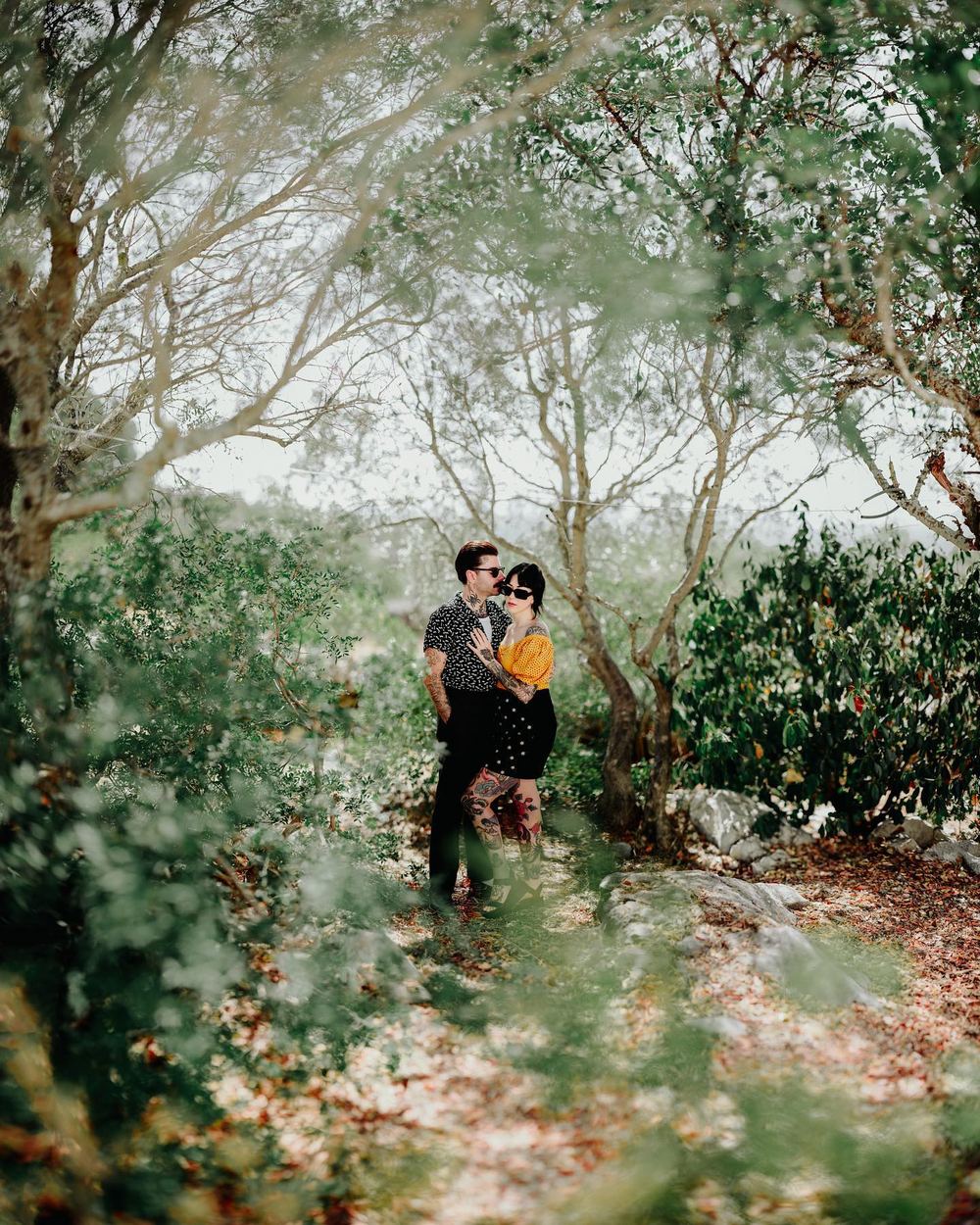 A couple holds each other among the trees. Seen through soft green foliage in bright daylight.