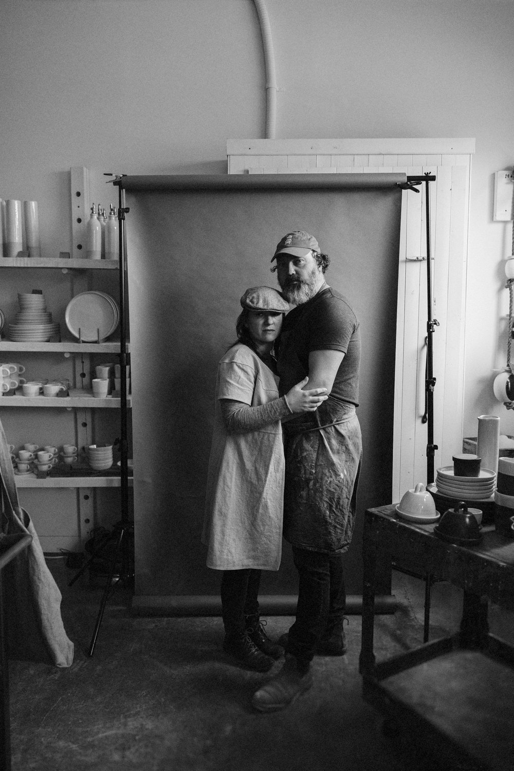 A couple stands close together in a pottery workshop before a plain backdrop. The man wears a clay stained apron. Shelves of plates, cups and ceramic pieces line the walls around them. Black and white.