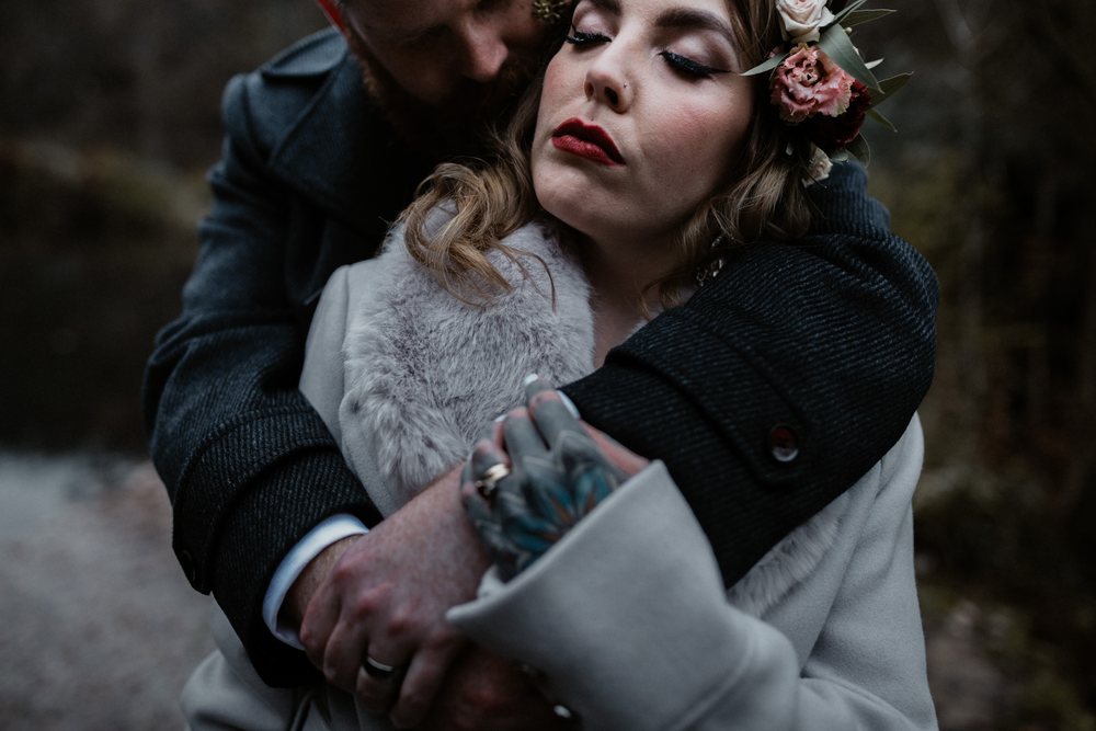 A bride wearing a crown of flowers rests with her eyes closed in the arms of her partner. A close embrace in soft winter light.